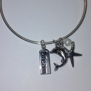 May include: A silver bangle bracelet with a silver dolphin charm, a silver starfish charm, a silver charm with the word "Dream" on it, and a white pearl charm.