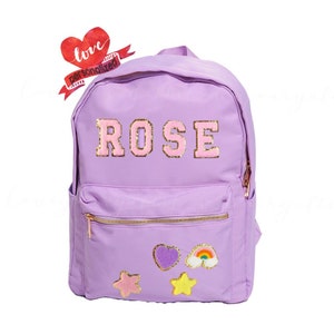 May include: Purple backpack with a gold zipper and the name "ROSE" in pink and gold letters. The backpack has a front pocket with a heart, rainbow, and star patches.