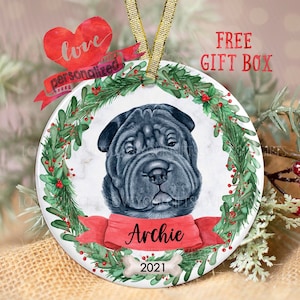 May include: A round, white ceramic Christmas ornament with a black Shar-Pei dog in a red bow. The ornament is decorated with a green wreath and red berries. The dog's name, "Archie", is written on the bow. The year "2021" is written on a bone below the bow.