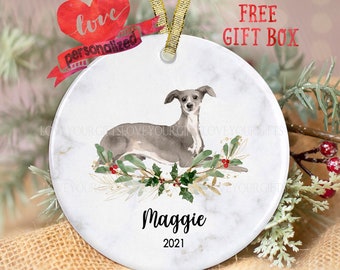 May include: A white ceramic Christmas ornament with a watercolor painting of a grey Italian Greyhound dog lying down on a bed of green holly and gold leaves. The ornament is personalized with the name "Maggie" and the year "2021".
