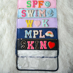 May include: A stack of ten colorful zippered pouches with different embroidered words and designs. The pouches are arranged vertically, with the top pouch reading "Beach" in pink letters and the bottom pouch reading "Skin" in pink letters with a red heart.