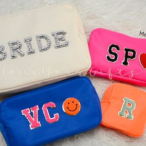 May include: Four different sizes of cosmetic bags with personalized lettering. The largest bag is cream colored with the word "BRIDE" in white pearl-like letters. The next largest bag is blue with the letters "VC" in pink. The medium bag is pink with the letters "SP" in black and a red heart. The smallest bag is orange with the letter "R" in gray.