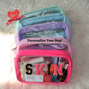 May include: Five clear plastic zippered pouches with a pink, purple, light blue, and green exterior. The pouches are stacked on top of each other. The bottom pouch has the word "SKIN" spelled out in large black letters with pink glitter. The text "Personalize Your Bag!" is printed on the pouch above the bottom pouch.