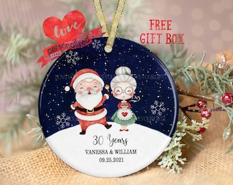 May include: A round, white, ceramic ornament with a blue background and a snowy scene. Santa Claus and a woman with a white beard are holding bells. The text "30 Years Vanessa & William 09.25.2021" is printed on the ornament.