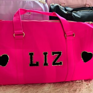 May include: A bright pink duffel bag with gold accents and black hearts. The bag has the name "LIZ" in black letters with gold trim.
