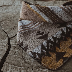 May include: A dog bandana with a geometric pattern in brown, beige, and white. The bandana is folded and displayed on a weathered wooden surface. The fabric has a textured appearance, and a small leather tag is attached. The design incorporates earthy tones.