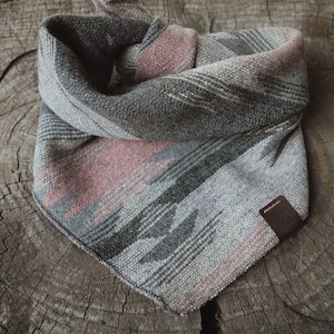 May include: A dog bandana in grey, pink, and black with a geometric pattern and a leather tag. The fabric has a woven texture. The bandana is knotted at the top and rests on a wooden surface.