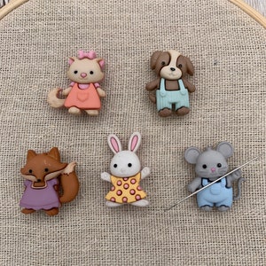 May include: Five colorful animal-shaped buttons with a needle and thread. The buttons are a brown fox, a white rabbit, a gray mouse, a brown and white dog, and a pink and white cat.
