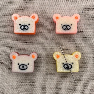 May include: Four small bear-shaped sponge erasers in different colors: orange, pink, yellow, and brown. The erasers are arranged in a square pattern on a beige fabric background.
