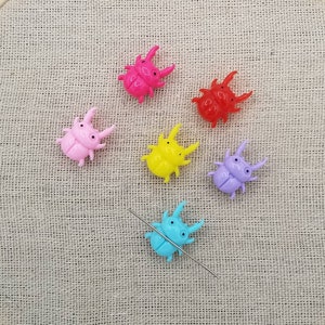 May include: A wooden embroidery hoop with beige fabric, holding six colorful plastic beetle-shaped embellishments. The beetles are pink, red, yellow, purple, and blue. A silver needle is also in the fabric.