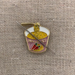 May include: A gold-coloured metal pin shaped like a cup of noodles with a red chilli pepper and the word "NOODLES" on the side. A silver needle is threaded through the pin.