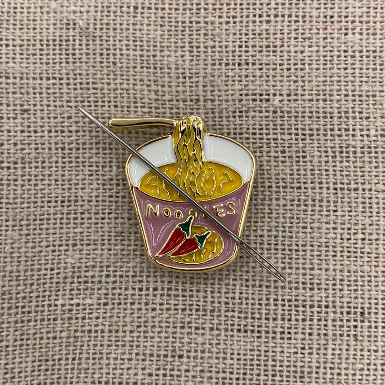 May include: A gold-coloured metal pin shaped like a cup of noodles with a red chilli pepper and the word "NOODLES" on the side. A silver needle is threaded through the pin.
