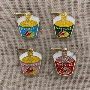 May include: Four gold-coloured enamel pins shaped like a bowl of noodles with a red chilli pepper. The pins say "NOODLES" in white letters.