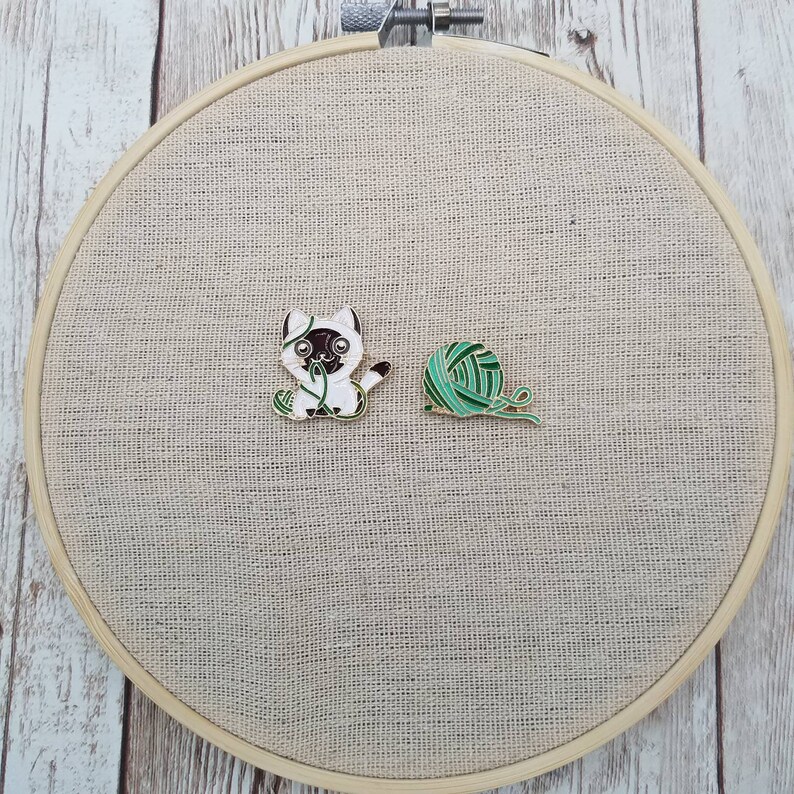 K&ouml;nnte beinhalten: Zwei Emaille-Pins auf einem beigefarbenen Stoffhintergrund. Ein Pin zeigt eine wei&szlig;e Katze mit schwarzen Markierungen, die eine gr&uuml;ne Wollkugel h&auml;lt. Der andere Pin ist eine gr&uuml;ne Wollkugel.