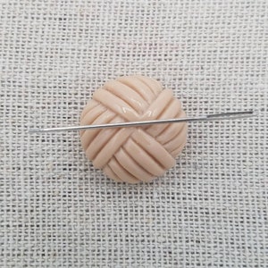 May include: A close-up of a beige button with a woven design. A needle is placed across the button.