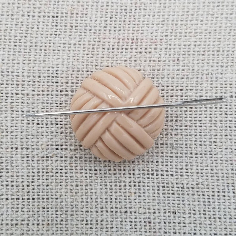 May include: A close-up of a beige button with a woven design. A needle is placed across the button.
