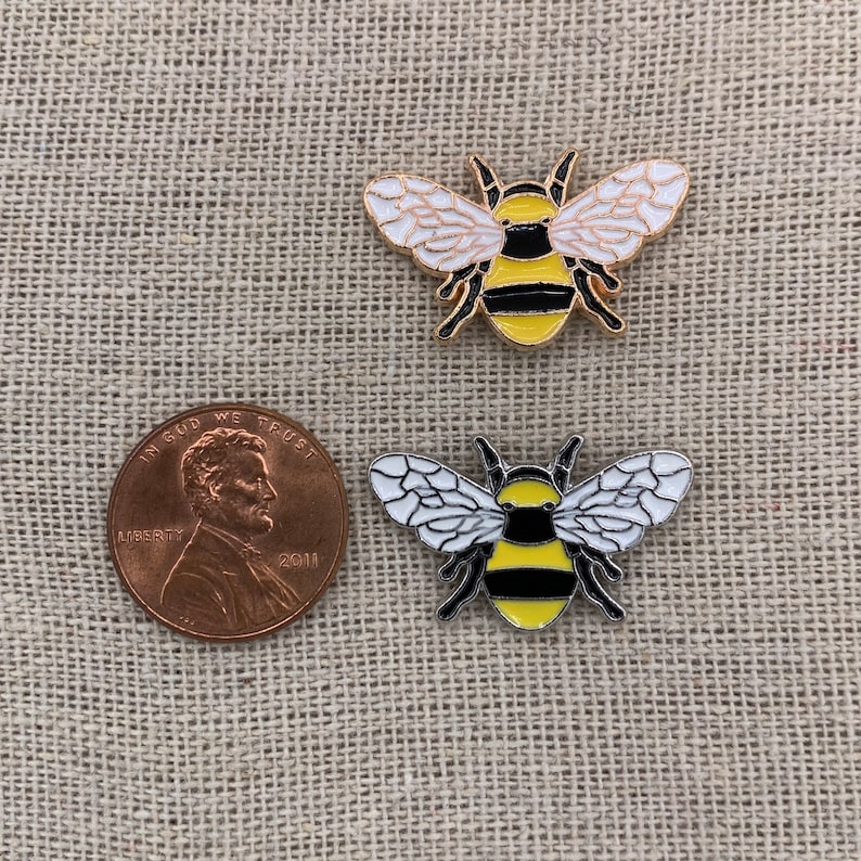 May include: Two gold-colored enamel pins shaped like bumblebees. The bees have black stripes, white wings, and yellow bodies. The pins are on a brown fabric background.