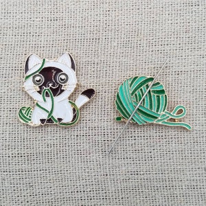 K&ouml;nnte beinhalten: Eine wei&szlig;e und braune Katze mit gr&uuml;nem Garn, eine gr&uuml;ne und goldene Garnkugel und eine silberne Nadel.