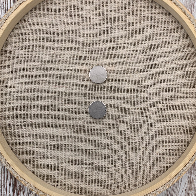May include: Two round silver magnetic buttons on a beige burlap background.