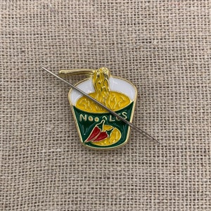 May include: A gold-coloured enamel pin shaped like a cup of noodles with a red chilli pepper inside. The pin has the word "NOODLES" written on the side. A silver needle is threaded through the pin.