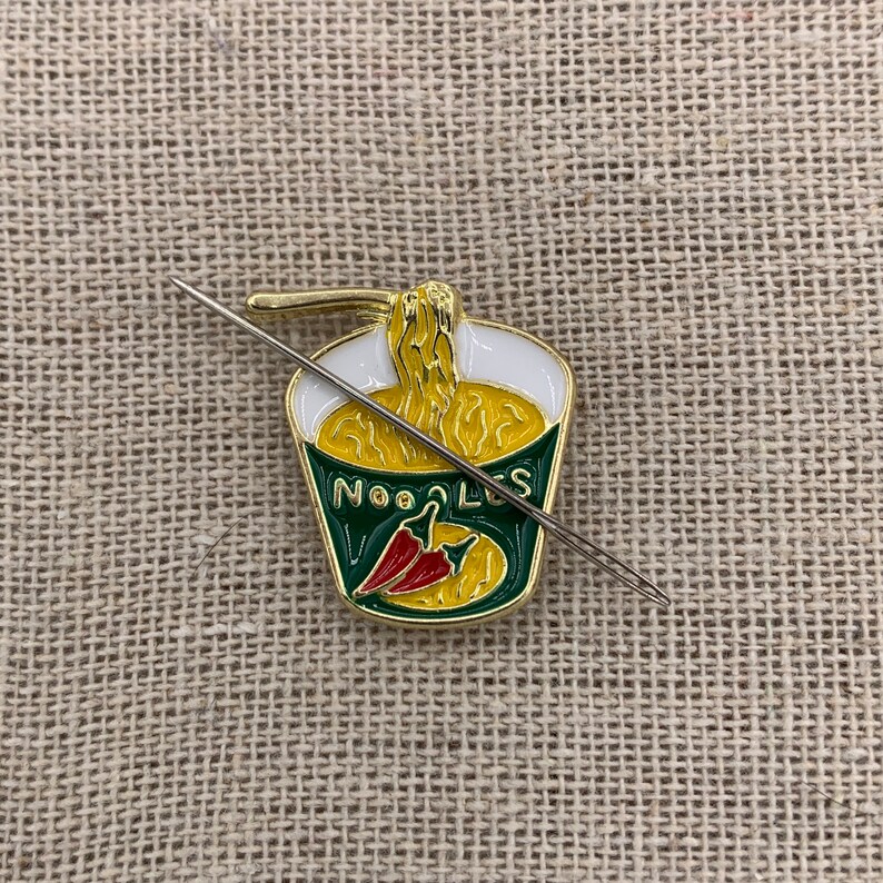 May include: A gold-coloured enamel pin shaped like a cup of noodles with a red chilli pepper inside. The pin has the word "NOODLES" written on the side. A silver needle is threaded through the pin.