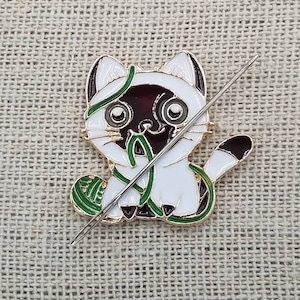 K&ouml;nnte beinhalten: Eine goldfarbene Metallstifte mit einem wei&szlig;en K&auml;tzchen mit braunen Ohren und einer schwarzen Nase. Die Katze h&auml;lt einen gr&uuml;nen Wollkn&auml;uel und eine Nadel geht durch die Wolle.