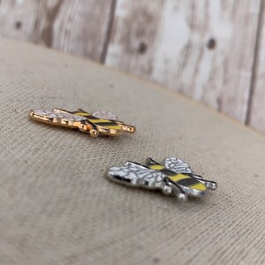 May include: Two enamel pins shaped like bees. One bee is gold with black stripes and the other is silver with black stripes. Both bees have white wings.