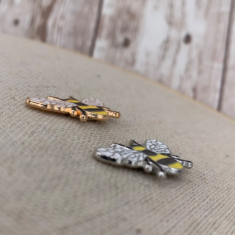 May include: Two enamel pins shaped like bees. One bee is gold with black stripes and the other is silver with black stripes. Both bees have white wings.