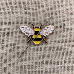 May include: A gold-colored bee pin with black stripes and white wings. The pin is pierced through the center with a needle.