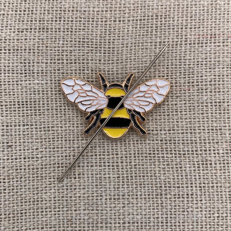May include: A gold-colored bee pin with black stripes and white wings. The pin is pierced through the center with a needle.