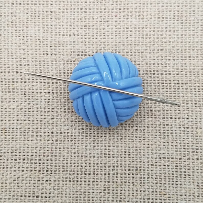 May include: A blue yarn ball with a silver knitting needle going through it. The yarn ball and needle are on a beige fabric background.