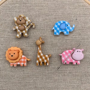 May include: A collection of animal-shaped embellishments, including a monkey, elephant, lion, giraffe, and hippo. Each animal has a gingham pattern in various colors. A needle is also present, suggesting a crafting theme.