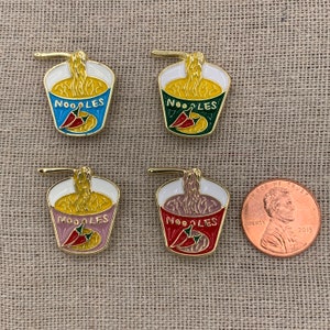 May include: Four enamel pins shaped like a cup of noodles with a red chili pepper and the word "NOODLES" on the front. The pins are in different colours: blue, green, pink, and red.