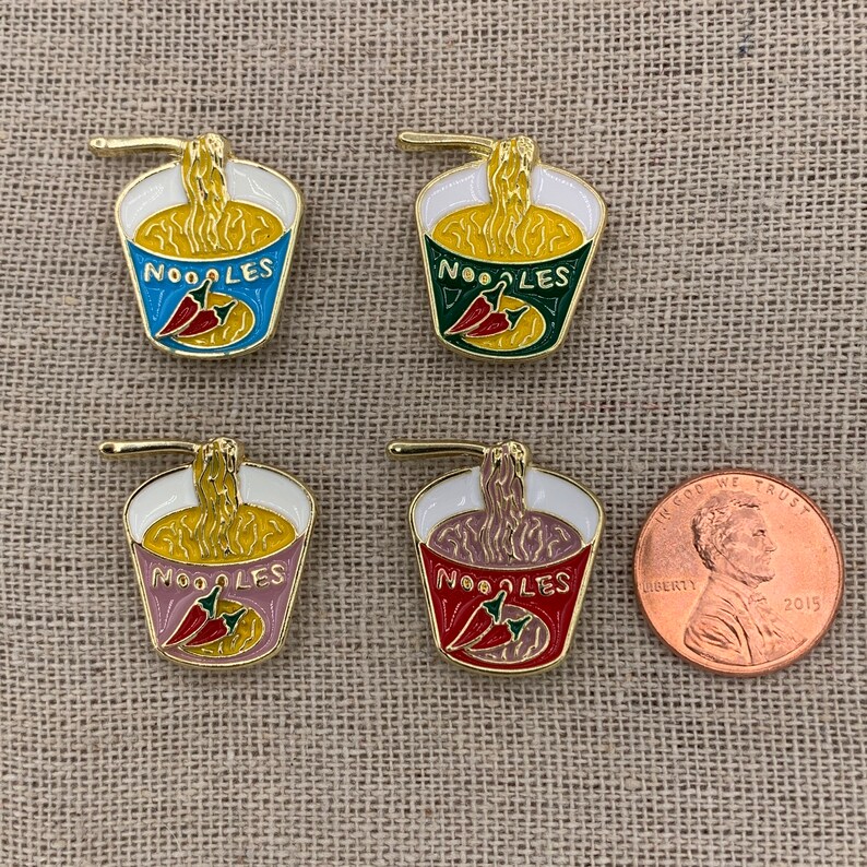 May include: Four enamel pins shaped like a cup of noodles with a red chili pepper and the word "NOODLES" on the front. The pins are in different colours: blue, green, pink, and red.