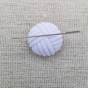 May include: A white plastic yarn ball with a needle going through it. The yarn ball is on a beige fabric background.