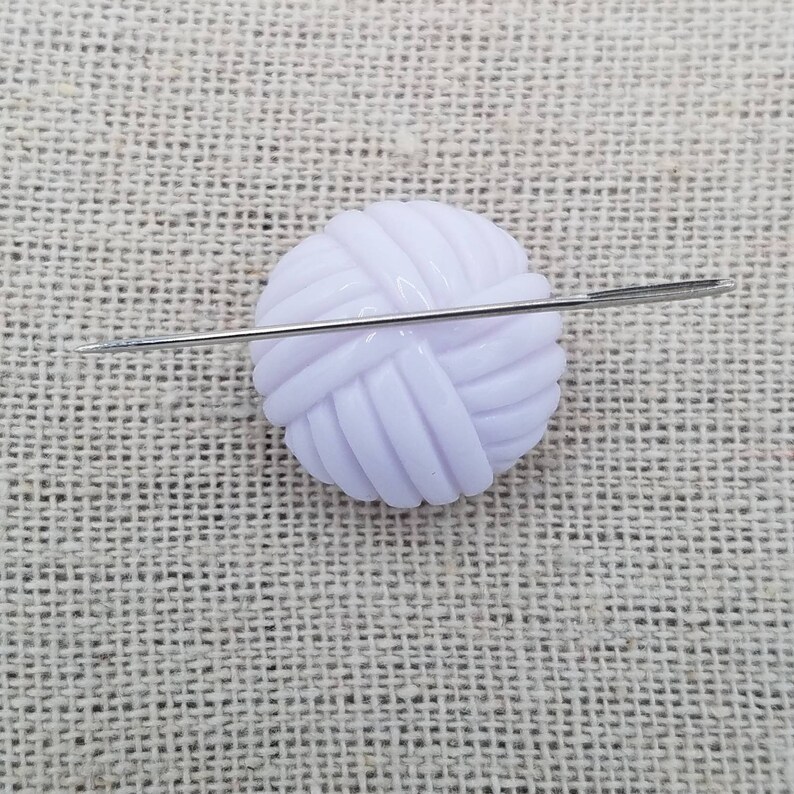 May include: A white plastic yarn ball with a needle going through it. The yarn ball is on a beige fabric background.