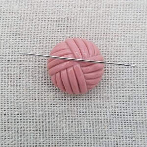 May include: A pink yarn ball with a silver knitting needle going through it. The yarn ball is on a light brown fabric background.