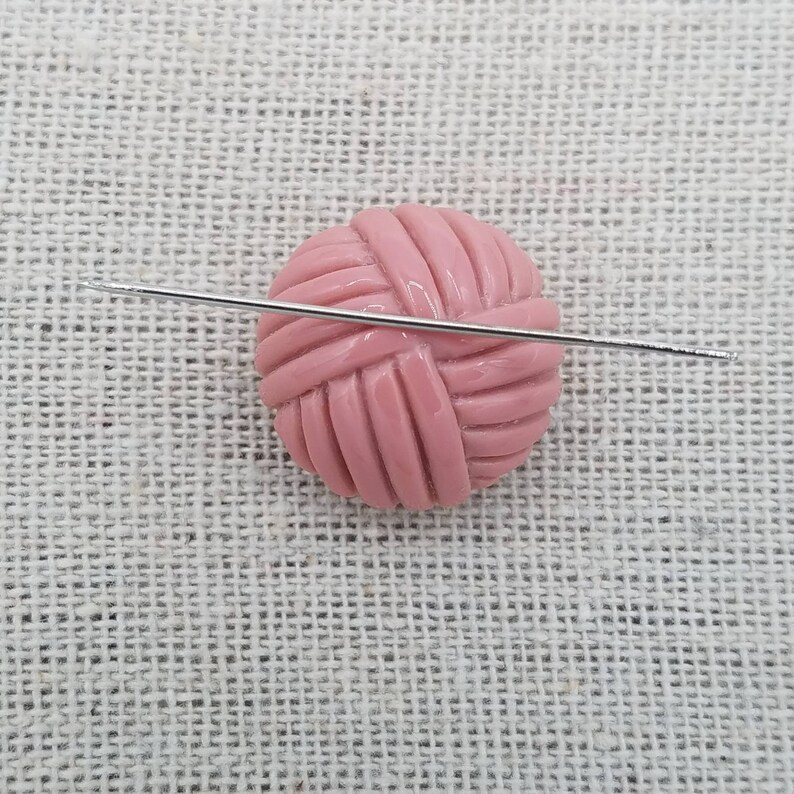 May include: A pink yarn ball with a silver knitting needle going through it. The yarn ball is on a light brown fabric background.