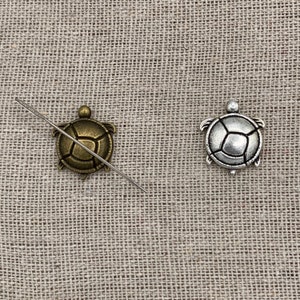 May include: Two metal turtle charms, one bronze and one silver, are displayed on a textured, neutral-toned surface. A needle is placed across the bronze turtle. The charms have detailed shell patterns and small flipper-like appendages.