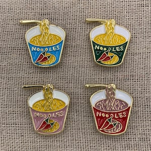 May include: Four enamel pins featuring a cartoon illustration of a cup of noodles with a red chili pepper. The pins are in four different colours: blue, green, pink, and red. Each pin says "NOODLES" on the cup.