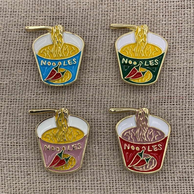 May include: Four enamel pins featuring a cartoon illustration of a cup of noodles with a red chili pepper. The pins are in four different colours: blue, green, pink, and red. Each pin says "NOODLES" on the cup.