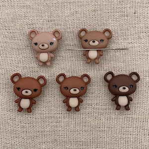May include: Five brown bear-shaped embellishments with varying shades of brown and tan. Each bear has a light tan belly, dark eyes, and small ears. The bears are arranged on a textured, beige surface.