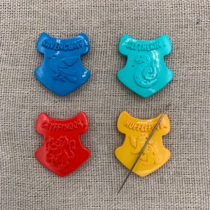 May include: Four colorful Harry Potter house crests: Ravenclaw (blue), Slytherin (teal), Gryffindor (red), and Hufflepuff (yellow). Each crest features a house name and emblem. A silver needle rests on the Hufflepuff crest. The background is a textured, neutral fabric.