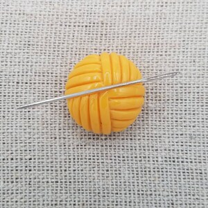 May include: A yellow yarn ball with a needle going through it. The yarn ball is on a beige fabric background.