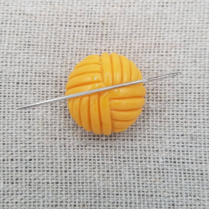 May include: A yellow yarn ball with a needle going through it. The yarn ball is on a beige fabric background.