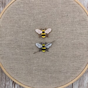 May include: Two bee pins on a beige burlap background. The bee on the top has gold wings and a black and yellow striped body. The bee on the bottom has white wings and a black and yellow striped body.