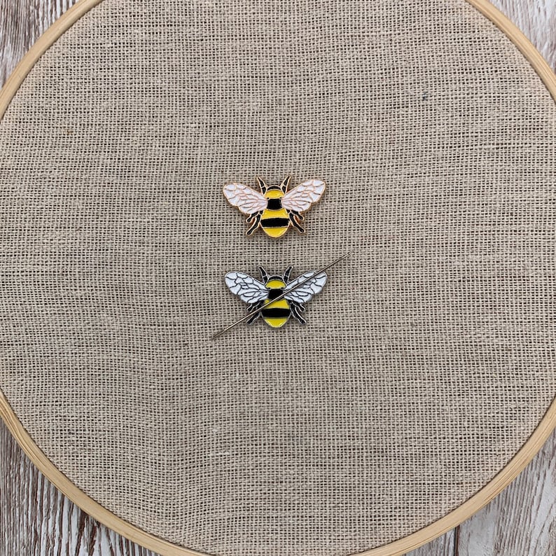 May include: Two bee pins on a beige burlap background. The bee on the top has gold wings and a black and yellow striped body. The bee on the bottom has white wings and a black and yellow striped body.