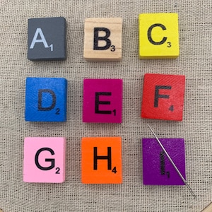 May include: Nine colorful wooden tiles with letters A through I and corresponding numbers. The tiles are arranged in three rows of three. The tiles are on a beige fabric background.