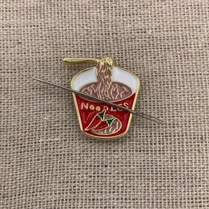 May include: A gold-coloured enamel pin of a red and white instant noodle cup with a noodle strand sticking out of the top. The cup says "NOODLES" and has two red chilli peppers inside. A silver needle is laying across the pin.