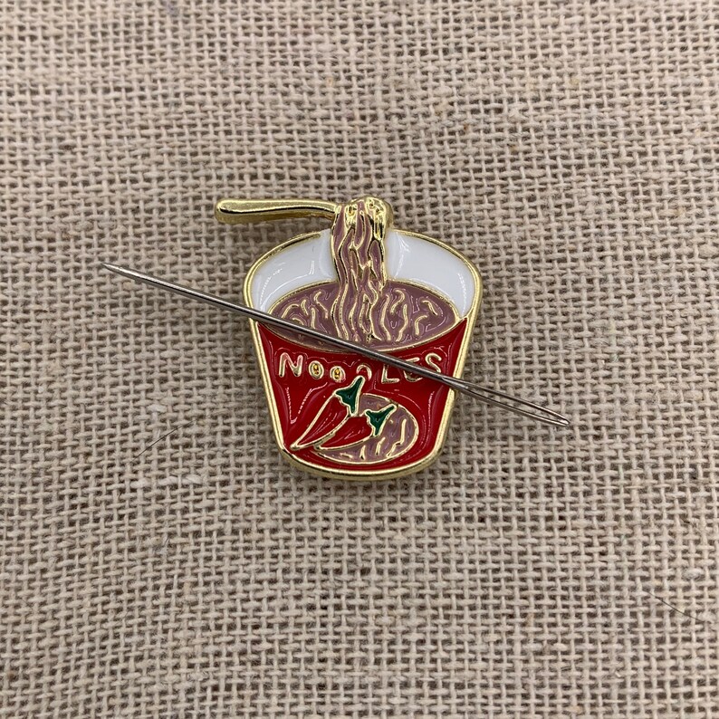 May include: A gold-coloured enamel pin of a red and white instant noodle cup with a noodle strand sticking out of the top. The cup says "NOODLES" and has two red chilli peppers inside. A silver needle is laying across the pin.